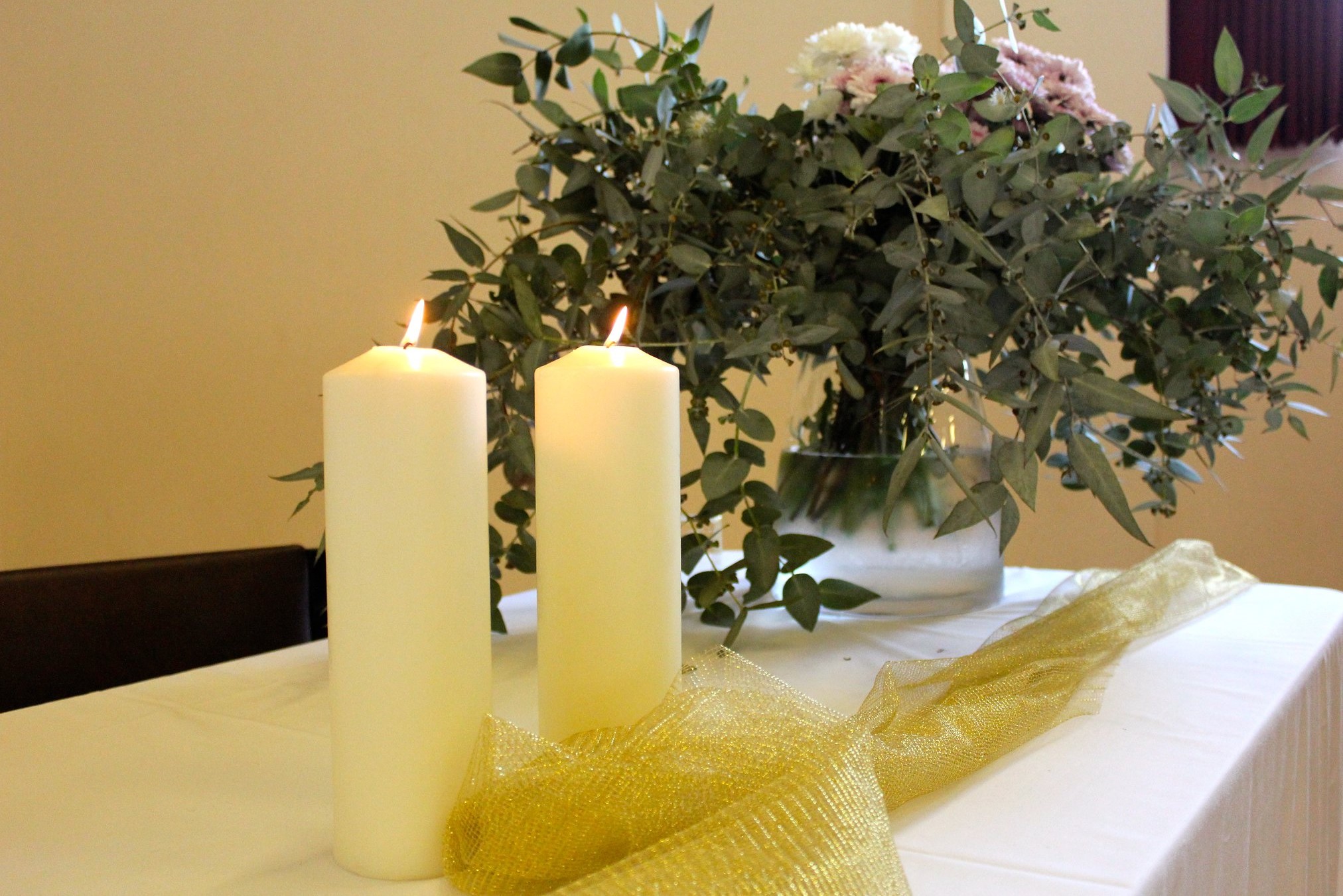 A picture of candles on a table covered with a white tablecloth with a vase of native flowers